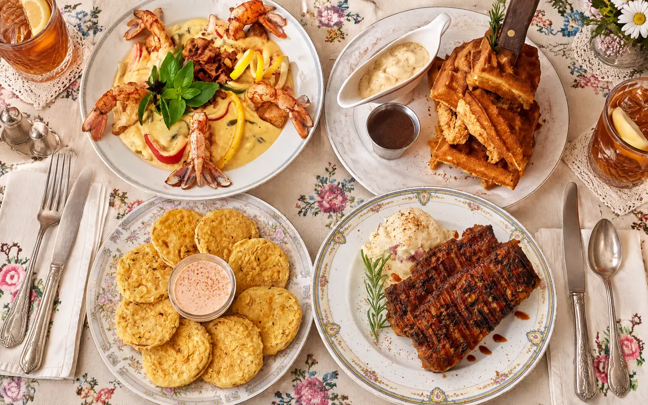 Four plates from the lunch and supper menu, seen from above: shrimp and grits, chicken and waffles with cream gravy, fried green tomatoes with remoulade, and glazed meatloaf over mashed potatoes, with glasses of sweet tea